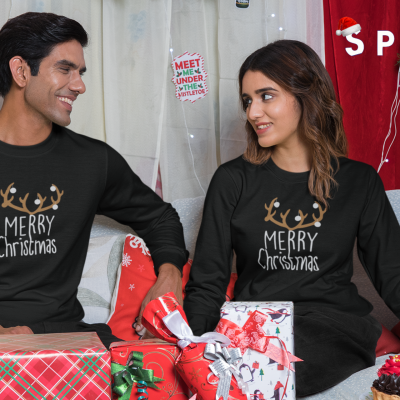 Homem e mulher vestidos com camisolas natalícias pretas, sentados com presentes de Natal ao redor em ambiente decorado para Natal.