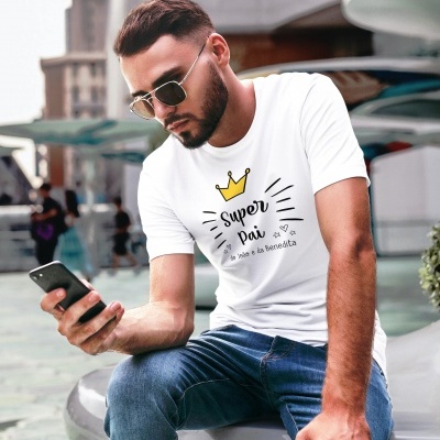 Homem com t-shirt branca com texto 'Super Pai a mãe e a Benedita' sentado em área urbana.