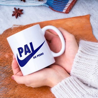 Caneca branca com texto 'PAI DO PEDRO' em azul escuro, segurada por mãos com camisola branca, sobre madeira e tecido cinzento.