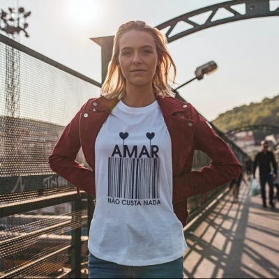 Mulher com camiseta branca com texto AMAR NÃO CUSTA NADA e casaco bordô numa passadeira ao ar livre