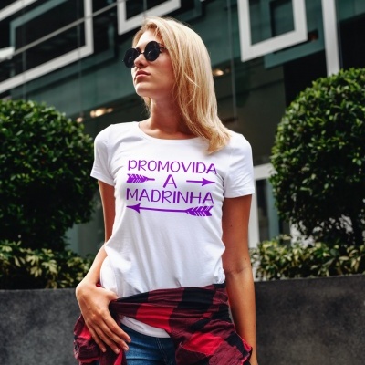 Mulher loira com t-shirt branca com texto roxo, óculos de sol e camisa xadrez presa à cintura em ambiente urbano ao ar livre.