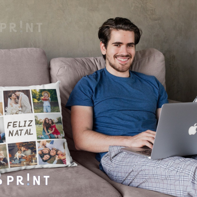 Almofada personalizada com fotos e texto FELIZ NATAL num sofá junto a homem com portátil.