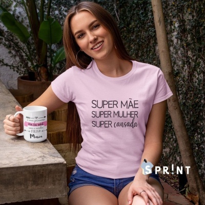 Mulher com camiseta rosa com texto 'SUPER MÃE SUPER MULHER SUPER cansada' e caneca branca em ambiente exterior