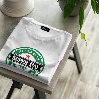 T-shirt branca dobrada com estampa 'FELIZ DIA DO PAI SUPER PAI' junto a uma planta em vaso