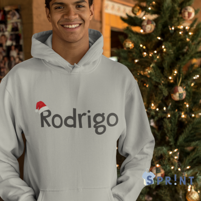 Homem a sorrir a usar camisola branca com capuz com nome 'Rodrigo' e gorro de Natal, árvore de Natal decorada atrás