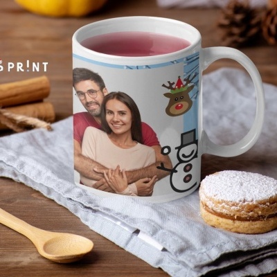 Caneca branca com fotografia de casal e desenhos natalícios, sobre pano cinza, com colher de madeira e doce ao lado.