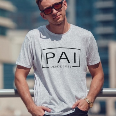 Homem com t-shirt cinza claro escrita PAI DESDE 2021 e óculos escuros em ambiente urbano
