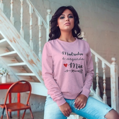 Mulher com sweatshirt rosa claro escrita Madrinha é segunda Mãe num ambiente interior com escada branca.