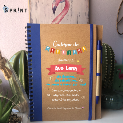 Caderno de receitas com capa decorada, elástico e caneta azul, sobre mesa com plantas e quadro flamingo ao fundo.