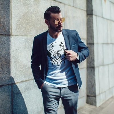 Homem de óculos de sol com t-shirt branca estampada e blazer azul junto a parede de pedra