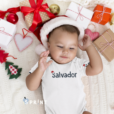 Bebé com body branco escrito Salvador e chapéu de Pai Natal rodeado de presentes e decorações natalinas