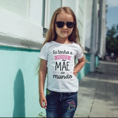 Criança a usar camiseta branca com texto em português e calças de ganga bordadas junto a uma parede azul e branca