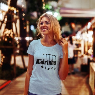 Mulher a sorrir usando t-shirt branca com texto 'Madrinha' num ambiente noturno iluminado