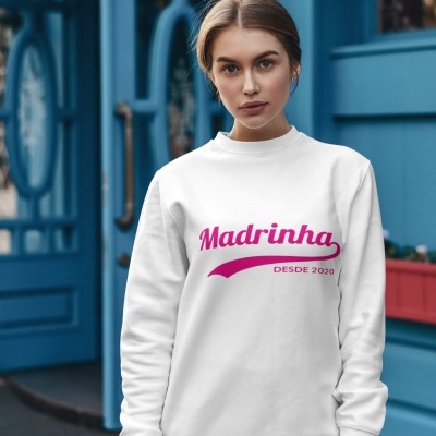 Mulher veste sweatshirt branca com texto rosa 'Madrinha DESDE 2020' em frente a porta azul