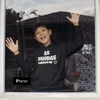 Rapaz de hoodie preto com texto branco 'AS MIÚDAS ADORAM-ME' encostado numa janela de vidro