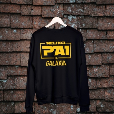 Casaco preto com texto amarelo 'MELHOR PAI DA GALÁXIA' pendurado em cabide de madeira contra parede de tijolos