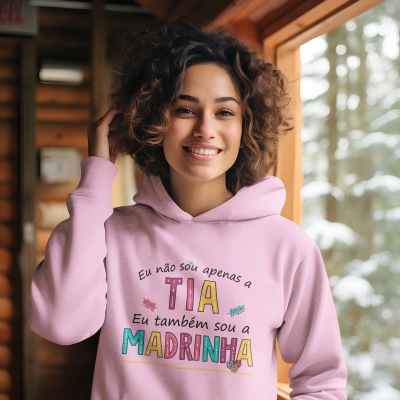 Mulher sorridente a usar suéter com capuz rosa claro com texto colorido, num ambiente de madeira junto a uma janela com neve lá fora