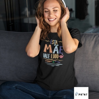 Mulher sorridente com camisola preta 'ESTA MÃE VALE 1.000 beijinhos do Pedro', sentada num sofá cinza escuro, calças de ganga e auscultadores brancos.