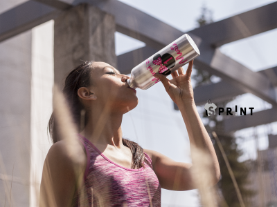 Mulher a beber água de uma garrafa metálica prateada com tampo vermelho ao ar livre