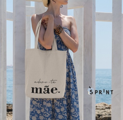 saco de tecido bege com texto adoro-te mãe pendurado no ombro de mulher com vestido azul floral junto ao mar