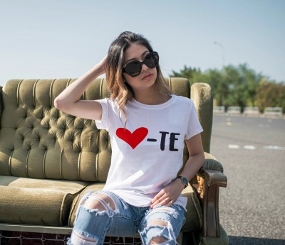 Mulher sentada num sofá verde usando t-shirt branca com estampa de coração vermelho e letras pretas