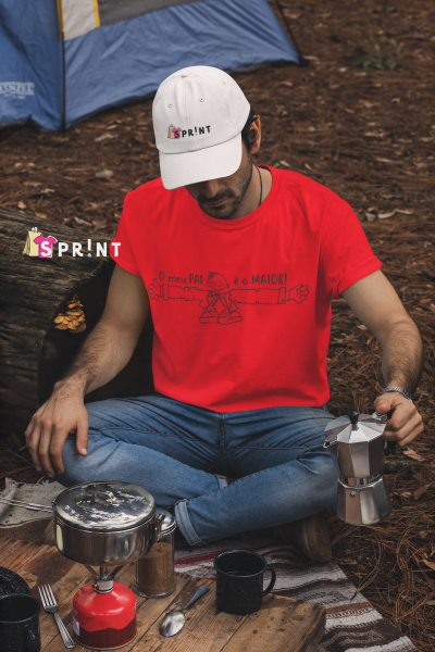 Homem com t-shirt vermelha e boné branco sentado no chão da floresta com utensílios de campismo.