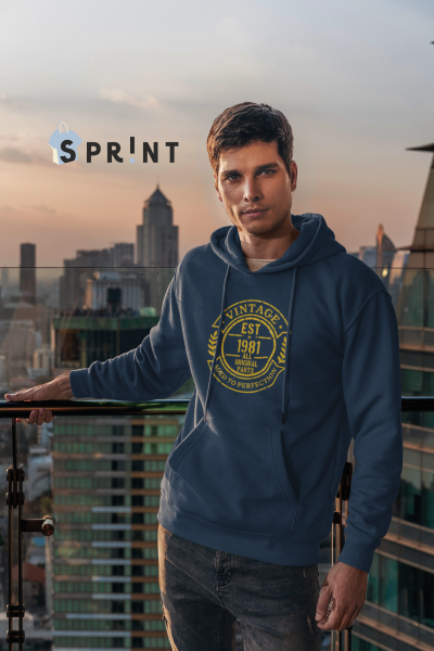 Homem jovem com casaco azul escuro com capuz e estampa amarela, em varanda com vista para cidade ao pôr do sol.