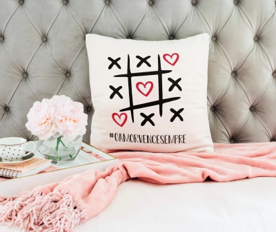 Capa de almofada branca com jogo do galo e corações vermelhos, texto '#OAMORVENCENSEMPRE', sobre cama com manta rosa e flores em vaso