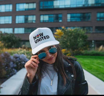 Mulher com boné branco NOW UNITED, óculos azuis e casaco preto em ambiente exterior