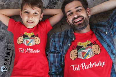 Rapaz e homem a usar t-shirts vermelhas com texto 'Tal Afilhado' e 'Tal Padrinho' e brasão, deitados no chão.