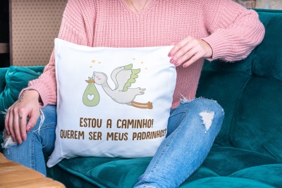 Travesseiro branco com estampa de cegonha e texto em castanho, segurado por pessoa sentada em sofá verde-azul.