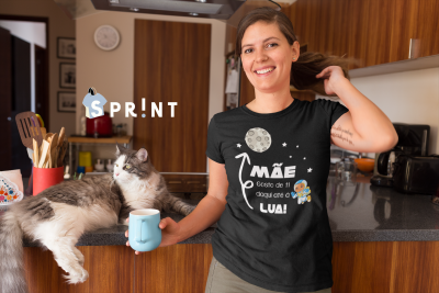 Camiseta preta com frase e ilustrações, usada por mulher numa cozinha com gato e caneca azul.