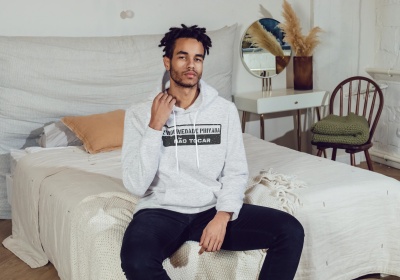 Homem sentado numa cama vestindo hoodie cinzento com texto preto e branco num quarto com decoração clara
