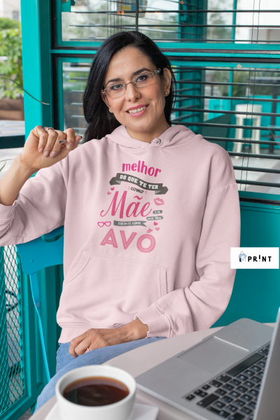 Mulher com sweat rosa e óculos sentada numa cadeira azul junto a mesa com computador e caneca de café