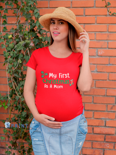 Mulher grávida com camisola vermelha com texto de Natal, chapéu de palha e saia de ganga, junto a uma parede de tijolos e plantas.