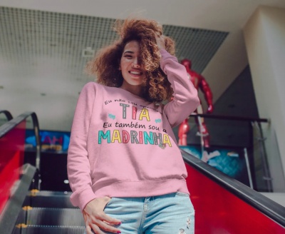 Mulher com sweatshirt rosa com texto colorido sobre ser tia e madrinha.