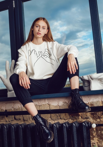 Moça loira em peitoril de janela com sweat branco, calças pretas e botas pretas