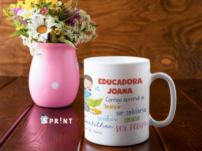 Caneca branca com texto colorido e jarro rosa com flores numa mesa de madeira.