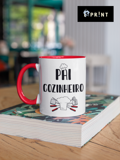 Caneca branca e vermelha com texto e desenho de utensílios de cozinha sobre livro em mesa.