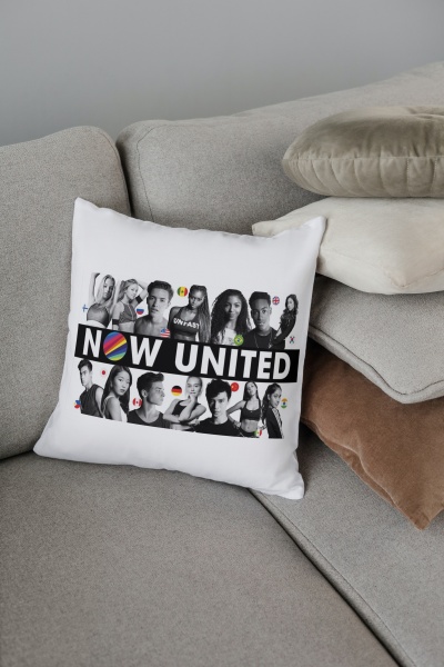 Almofada branca com imagem e texto 'NOW UNITED' sobre sofá cinzento, com outras almofadas de cores neutras ao lado.