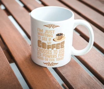 Caneca branca com texto alusivo ao café e uma chávena de café desenhada, sobre superfície de madeira.