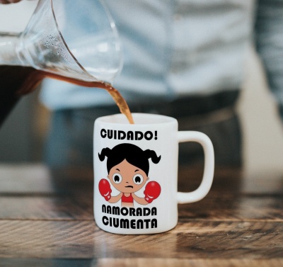 Caneca branca com desenho de menina e texto CUIDADO! NAMORADA CIUMENTA, bebida a ser derramada nela.