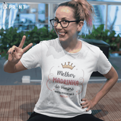 Pessoa com t-shirt branca com texto sobre madrinha, óculos e cabelo apanhado, fundo interior com plantas e objetos desfocados