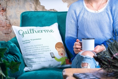 Almofada com texto explicativo do nome Guilherme num sofá azul tejido, pessoa segurando caneca ao lado