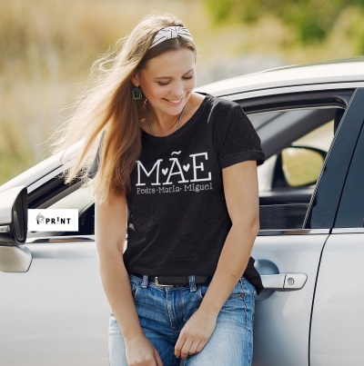 Mulher com t-shirt preta com texto MÃE e nomes, ao lado de carro prata
