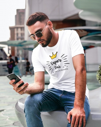 Homem com t-shirt branca com texto 'Super Pai a mãe e a Benedita' sentado em área urbana.