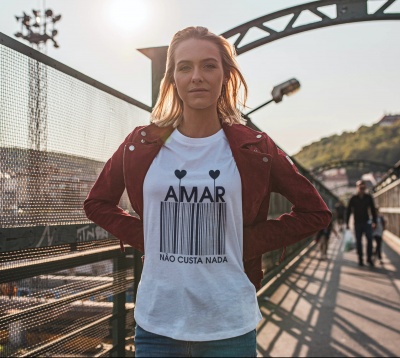 Mulher com camiseta branca com texto AMAR NÃO CUSTA NADA e casaco bordô numa passadeira ao ar livre