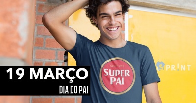 Homem sorridente com camisola azul 'SUPER PAI' em fundo amarelo e parede de tijolos vermelhos