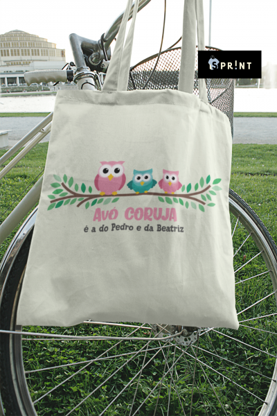 Saco de pano bege com corujas coloridas e texto pendurado num guiador de bicicleta ao ar livre