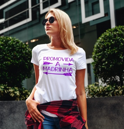 Mulher loira com t-shirt branca com texto roxo, óculos de sol e camisa xadrez presa à cintura em ambiente urbano ao ar livre.